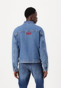 Chaqueta de mezclilla azul con cierre de botones en el frente, bolsillos laterales y un logo bordado en rojo de 'Red Bull Racing' en la parte posterior.