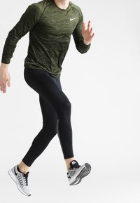 Träningsoutfit bestående av en grön, strukturerad långärmad tröja, svarta leggings och svarta/vita löparskor med ett mönstrat sula.