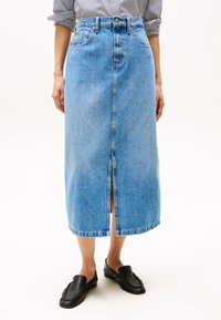 Helles blaues Jeans-Midijugend mit einem Schlitz vorne, Gürtelschlaufen und zwei vorderen Taschen. Getragen mit schwarzen Slippern.