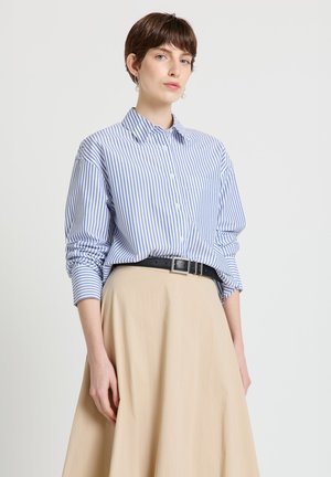 Woman wearing blue and white striped button-up shirt tucked into beige skirt with black belt, standing against a plain white background.