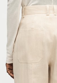 Κοντινό πλάνο μπεζ παντελονιού ψηλής μέσης με πίσω τσέπη και θηλιές για ζώνη, φορεμένο με ανοιχτό μπεζ μακρυμάνικη μπλούζα σε άτομο με σκουρόχρωμο δέρμα.