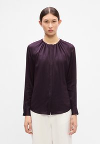 Dunkellila Seidenbluse mit gerafftem Ausschnitt, langen Ärmeln, vorderen Falten und abgerundetem Saum, kombiniert mit cremefarbenen Hosen.