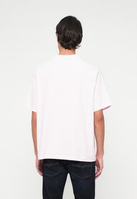 Mann mit dunklen Haaren, der ein schlichtes, weißes, übergroßes T-Shirt mit kurzen Ärmeln und eine dunkelblaue Jeans trägt, mit dem Rücken zu einer weißen Wand.