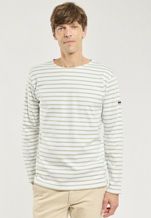 Homme aux cheveux châtain clair portant une chemise blanche à manches longues avec des rayures horizontales beige et un pantalon beige, debout devant un fond uni.