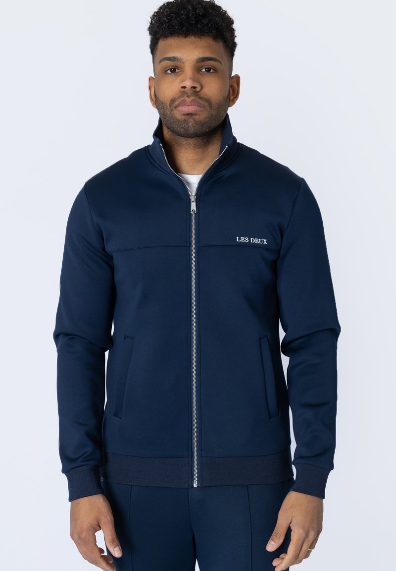 Jachetă zip-up de culoare bleumarin, realizată dintr-un material neted, având un guler înalt, buzunare laterale și broderie discretă a logo-ului pe piept.