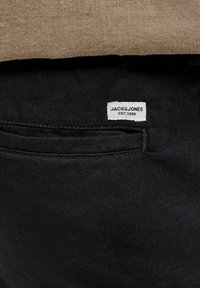 Μαύρο ύφασμα με μια μικρή λευκή ετικέτα της μάρκας Jack & Jones ραμμένη κοντά σε οριζόντιο άνοιγμα τσέπης.