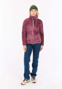 Fleece jednofarebná bunda na zips v malinovej farbe s vysokým golierom a detailom na šnúrke. Kombinovaná s modrými džínsami a béžovými teniskami. Na hlave má olivovozelenú čiapku.