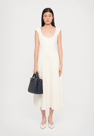 Sleeveless, cream-colored dress with a scoop neckline, fitted bodice, and A-line skirt. Model holds a dark navy handbag. Shoes are white with a pointed toe.
