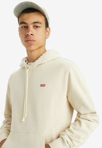 Cream-colored hoodie made of soft fabric, featuring a kangaroo pocket, adjustable drawstring hood, and small red Levi's logo on the chest.