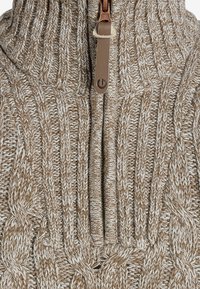 Hellbrauner gestrickter Pullover mit einem groben Kabelmuster. Verfügt über einen hohen Kragen und einen Reißverschluss bis zur Hälfte mit einem Leder-Zipp-Anhänger.