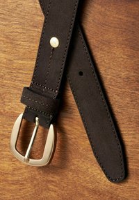 Ceinture en daim marron avec une boucle en métal doré, présentant des coutures contrastantes et un détail de rivet en métal. Le tout sur un fond en bois.