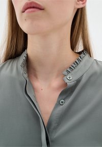 Camicia button-up grigia chiara con tessuto strutturato, caratterizzata da un colletto arricciato e due chiusure a bottone all'apertura del collo.