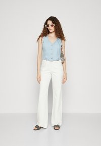 Světle modrý, bezrukávový džínový crop top se zapínáním na knoflíky, spárovaný s bílými širokými kalhotami. Doplňky zahrnují texturovanou tašku a sluneční brýle.