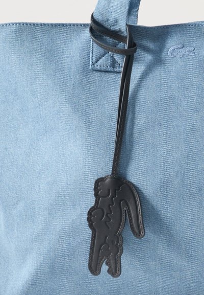 Gros plan sur un sac en denim bleu clair, orné d’un charm en cuir noir en forme de crocodile, suspendu à sa bandoulière.