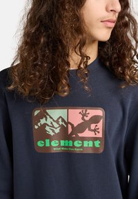 Granatowy sweter z grafiką przedstawiającą góry i jaszczurkę, z napisem "element" w kolorze zielonym oraz "Wind Water Fire Earth" poniżej.