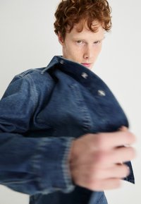 Giubbotto di jeans, blu scuro, vestibilità rilassata. Presenta bottoni a pressione metallici, colletto classico e dettagli di cucitura. Il tessuto ha una consistenza morbida.