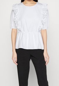 Weiße Bluse mit Rüschensleeves und bestickten Details, mit taillierter Taille und lässigem Schnitt. Dazu schwarze Hosen.