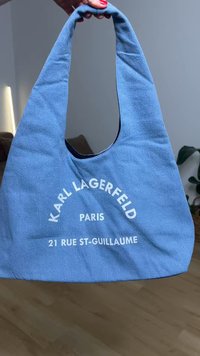 Hånd som holder en myk blå stoffpose med "KARL LAGERFELD PARIS 21 RUE ST-GUILLAUME" trykt i hvitt på forsiden.