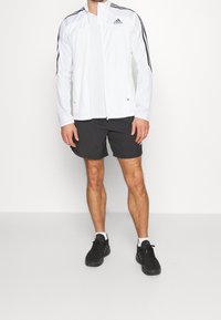 Vit idrottjacka med svarta ränder, dragkedja framtill, ihop med svarta shorts. Svarta löparskor med vita detaljer.