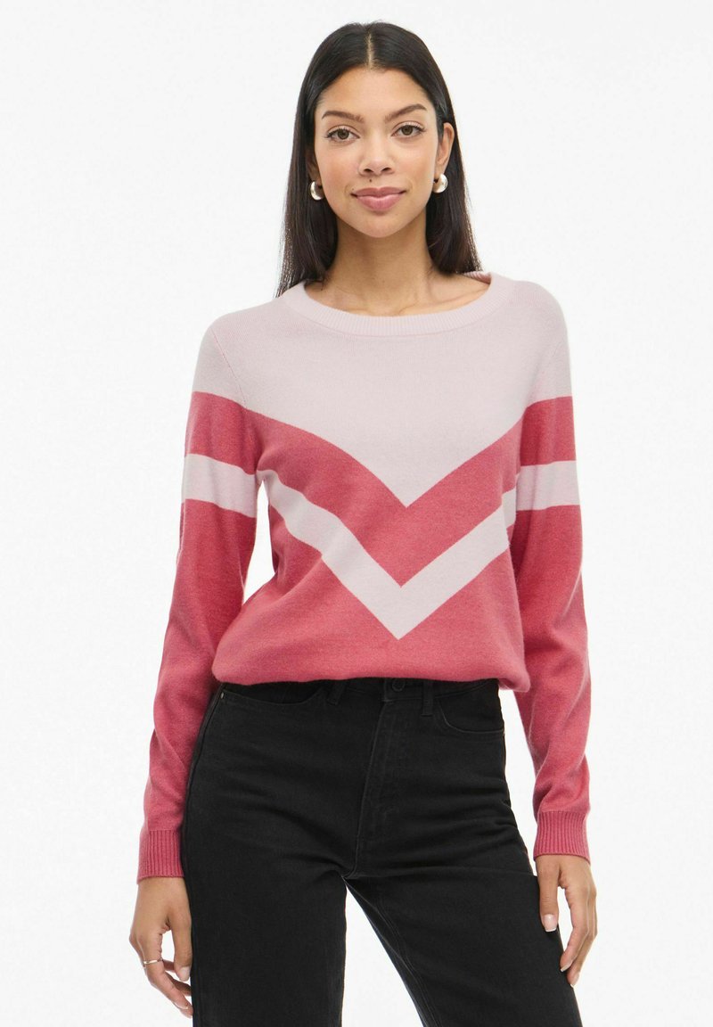 Sweter dzianinowy z wzorem w zygzak w odcieniach różu i czerwieni, okrągły dekolt i długie rękawy, w połączeniu z czarnymi dżinsami.
