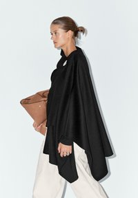 Poncho nero, di tessuto testurizzato, con colletto alto e forma asimmetrica. Abbinato a pantaloni leggeri e larghi e a una borsa a tracolla in pelle marrone.