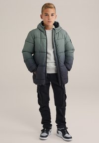 Puffjacka i gradientgrön till svart, med huva, quiltat mönster, två sidofickor, tillsammans med svarta cargo-byxor och sneakers.