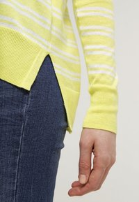 Pull à manches longues rayé jaune et blanc avec une fente latérale, porté avec un jean bleu, montrant l'avant-bras et la main.