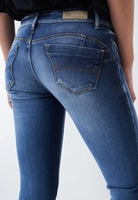 Vaqueros de mezclilla azul oscuro con corte ajustado, que cuentan con una cintura de altura media, bolsillos traseros con detalles de costura y un ligero desgaste en la tela.