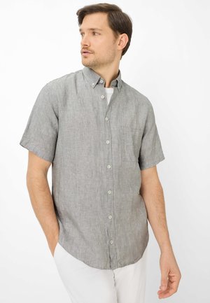 Mann trägt ein kurzärmeliges, graues Hemd mit Knöpfen und Brusttasche, steht mit einer Hand in der Tasche und schaut zur Seite.