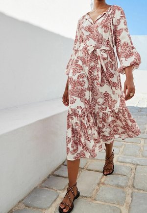 Frau, die auf einem Kopfsteinpflasterweg geht, trägt ein weißes und rostbraunes Paisleymuster-Kleid mit Bindegürtel und schwarze Nieten-Sandalen.