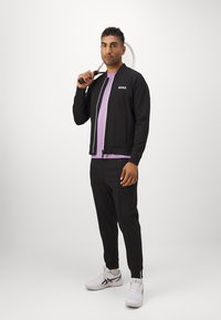 Svart zip-up jacka med "BOSS"-logotyp, tillsammans med en ljuslila skjorta och svarta byxor. Bär vita träningsskor och håller en tennisracket.