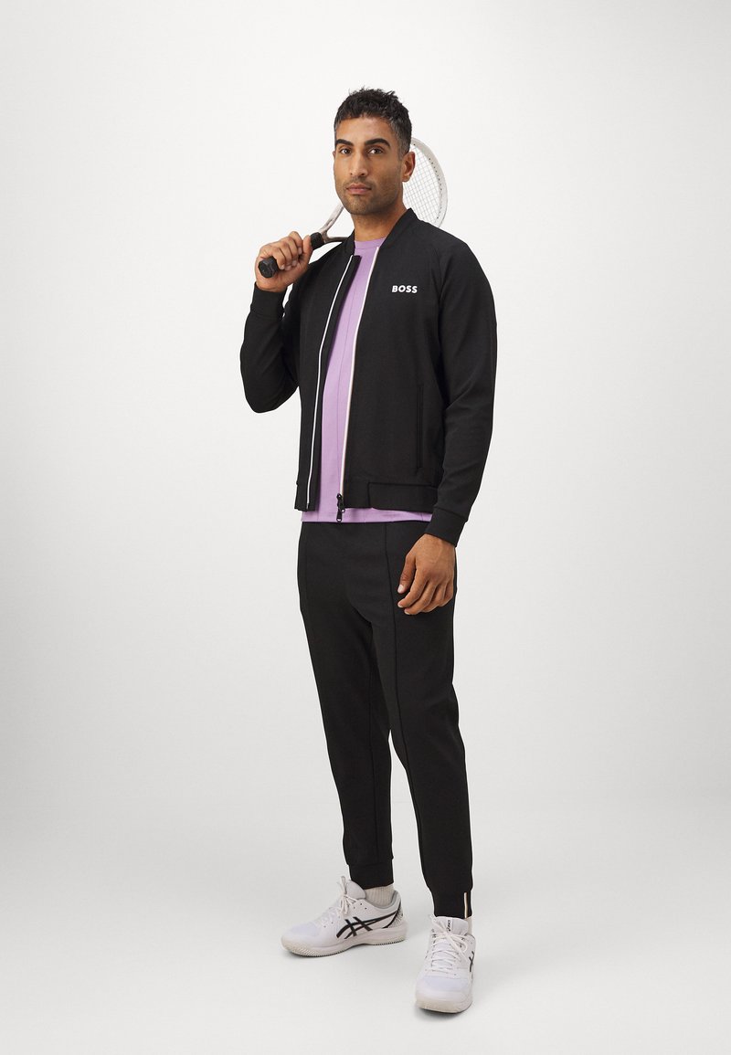 Svart zip-up jacka med "BOSS"-logotyp, tillsammans med en ljuslila skjorta och svarta byxor. Bär vita träningsskor och håller en tennisracket.
