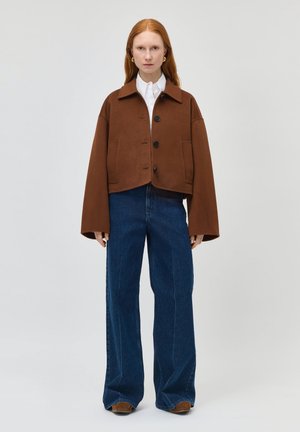 Kobieta z rudymi włosami ubrana w brązową, oversize zapinaną na guziki kurtkę, białą koszulę, szerokie niebieskie jeansy i brązowe buty, stojąca na jednolitym tle.