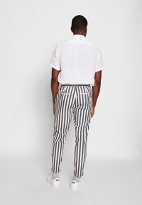 Man die wegkijkt en een wit T-shirt met korte mouwen draagt, zwart-witte verticaal gestreepte broek, grijze sokken en witte sneakers.