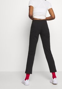 Person med svarta byxor, vit kortärmad T-shirt, röda strumpor och vita sneakers, stående vänd mot en vit vägg med händerna bakom ryggen.