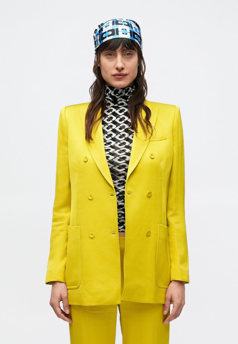 Blazer doppiopetto giallo brillante abbinato a un dolcevita bianco e nero con motivo a stampa, completato da un foulard per la testa con stampa geometrica blu.