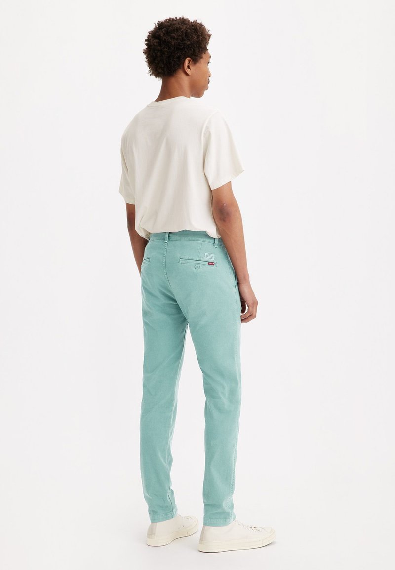 Pantalones de corduroy verde claro con un ajuste ceñido, bolsillos traseros y una pequeña etiqueta. Combinados con una camisa de manga corta color crema y zapatillas blancas.