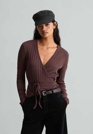 Jonge vrouw die een bruine wikkeltrui draagt, zwarte ribfluwelen broek met riem en een zwarte platte pet, staand met handen in de zakken.