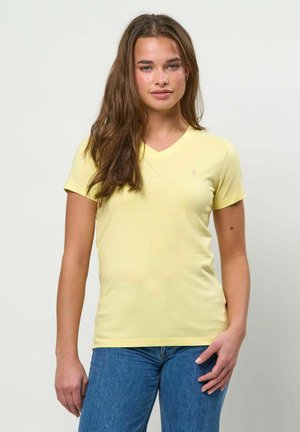 Tânără cu păr lung și castaniu, purtând un tricou galben deschis cu decolteu în V și blugi albaștri, stând în picioare pe un fundal alb simplu.