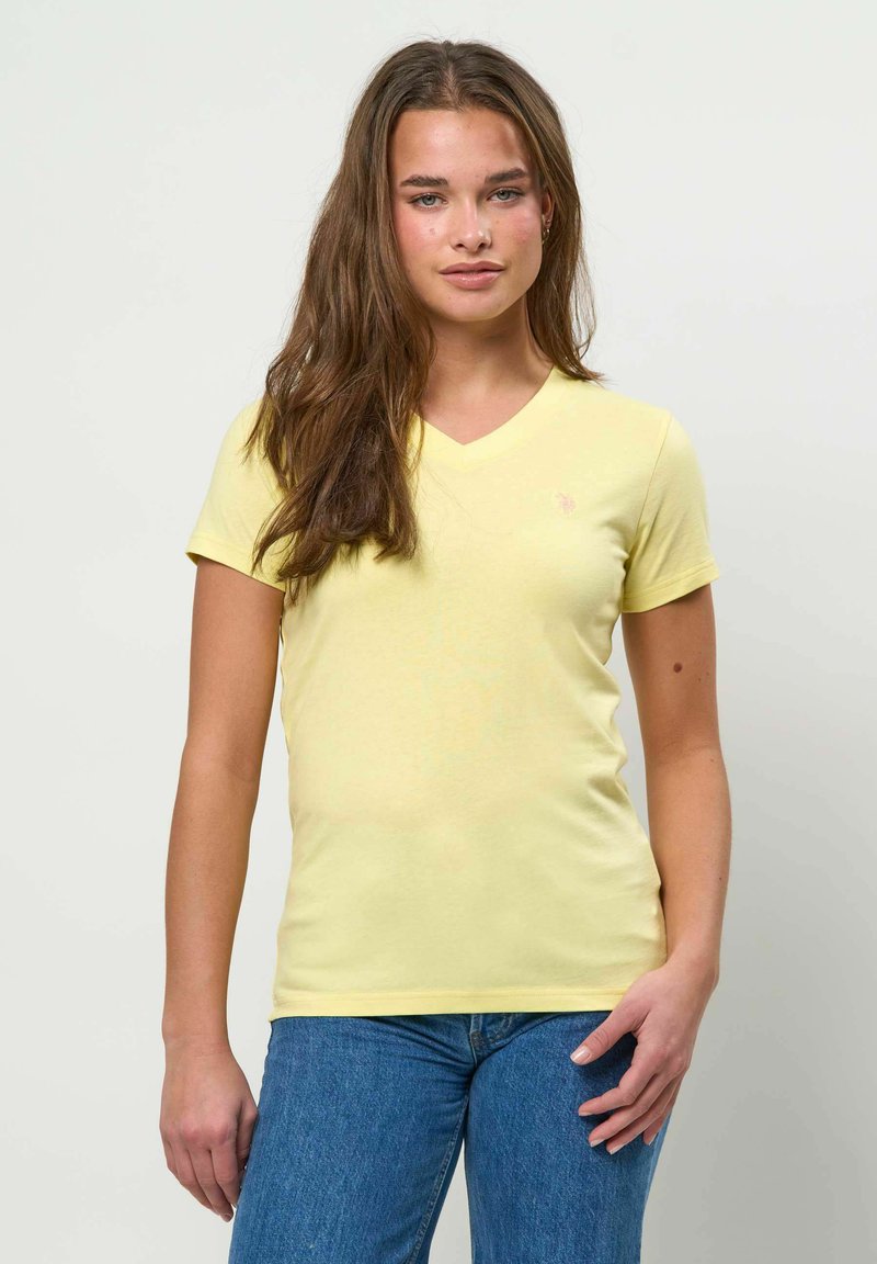 Jeune femme aux longs cheveux bruns portant un T-shirt jaune pâle à encolure en V et un jean bleu, debout devant un fond blanc uni.