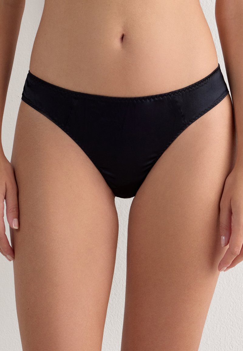 Kvinde iført sort satin bikini, der viser talje, hofter og øvre lår mod en ensfarvet lys baggrund.