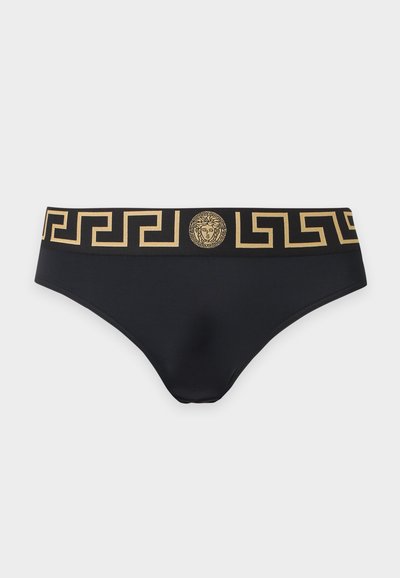 Slip noir avec une ceinture dorée à motif grec et un emblème doré de tête de Méduse centré à l'avant. Aucun objet visible à l'arrière-plan.