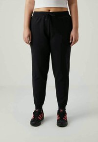 Άτομο που φοράει μαύρο παντελόνι jogger με κορδόνι στη μέση, λευκή κοντή μπλούζα και μαύρα με κόκκινες αθλητικές ακριβώς, που στέκεται σε ελαφρώς ανοιχτό φόντο.