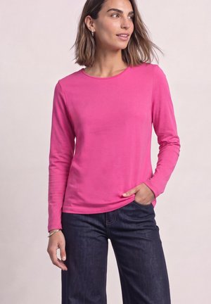 Kvinde iført en knaldrød langærmet skjorte og mørkeblå jeans, står med den ene hånd i lommen og kigger til siden.