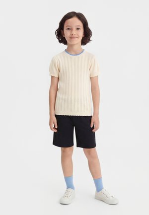 Bambino con capelli corti e ricci indossa una maglia lavorata a maglia crema a maniche corte, pantaloncini neri, calzini blu e scarpe da ginnastica bianche, in piedi contro uno sfondo bianco.
