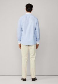 Uomo visto di spalle che indossa una camicia light blue con polsini bianchi, pantaloni beige e scarpe marroni scure su uno sfondo neutro.