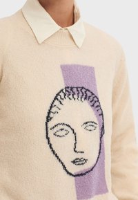 Knit sweater in cream with a graphic face design in black and purple. Features a contrasting light-colored collared shirt underneath.