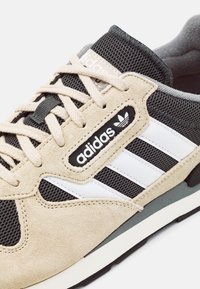 Zapatilla de ante beige y malla negra con rayas blancas, logo negro y detalles en gris. Presenta cordones ligeros y un cuello acolchado.