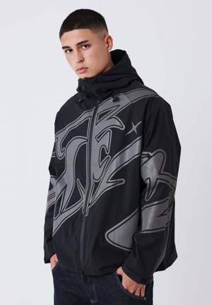 Jeune homme aux cheveux courts portant une veste noire à capuche avec de grands motifs graphiques gris et les mains dans les poches.