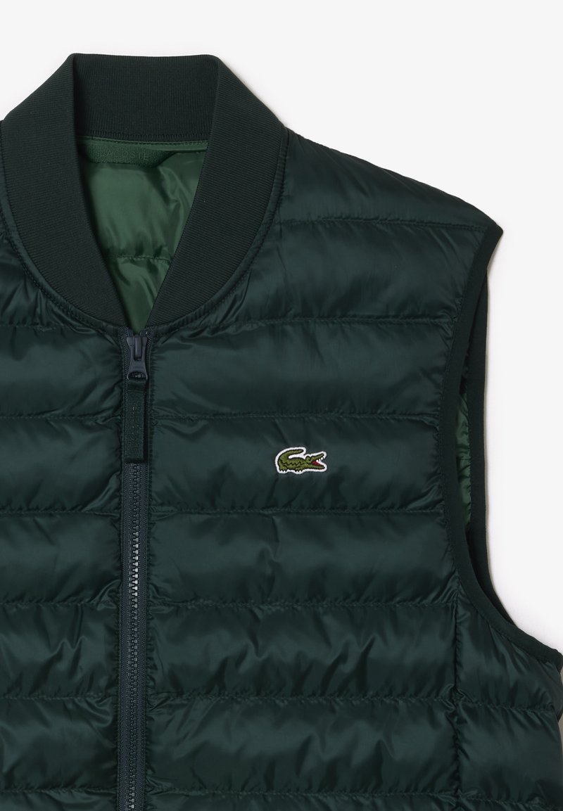 Chaleco acolchado verde oscuro con cuello acanalado, cierre frontal de cremallera y un logo bordado de cocodrilo en el lado izquierdo. Textura suave.